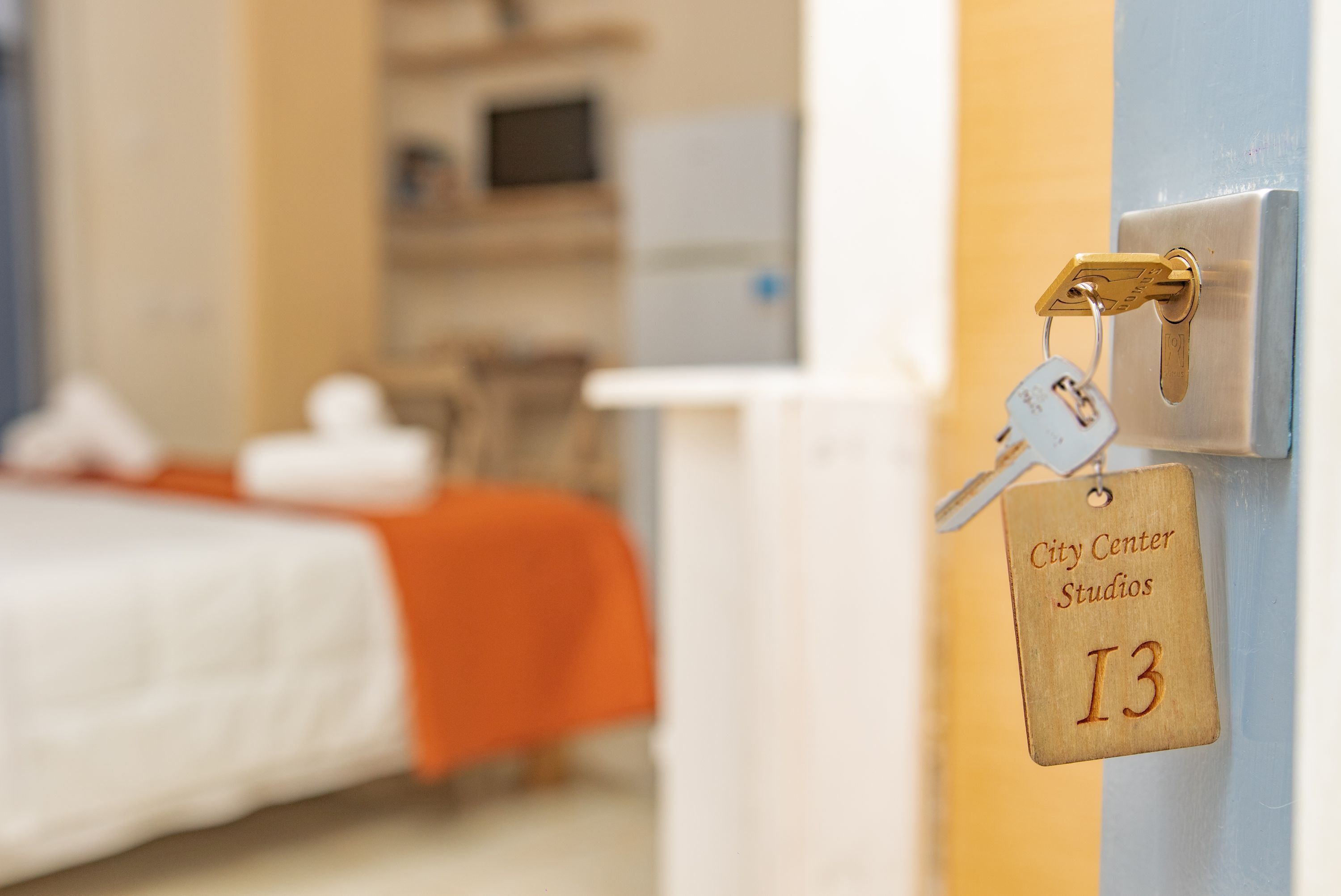 A hotel room with a bed, an orange blanket, a keychain with a tag reading "City Center Studios I3", and a door lock.