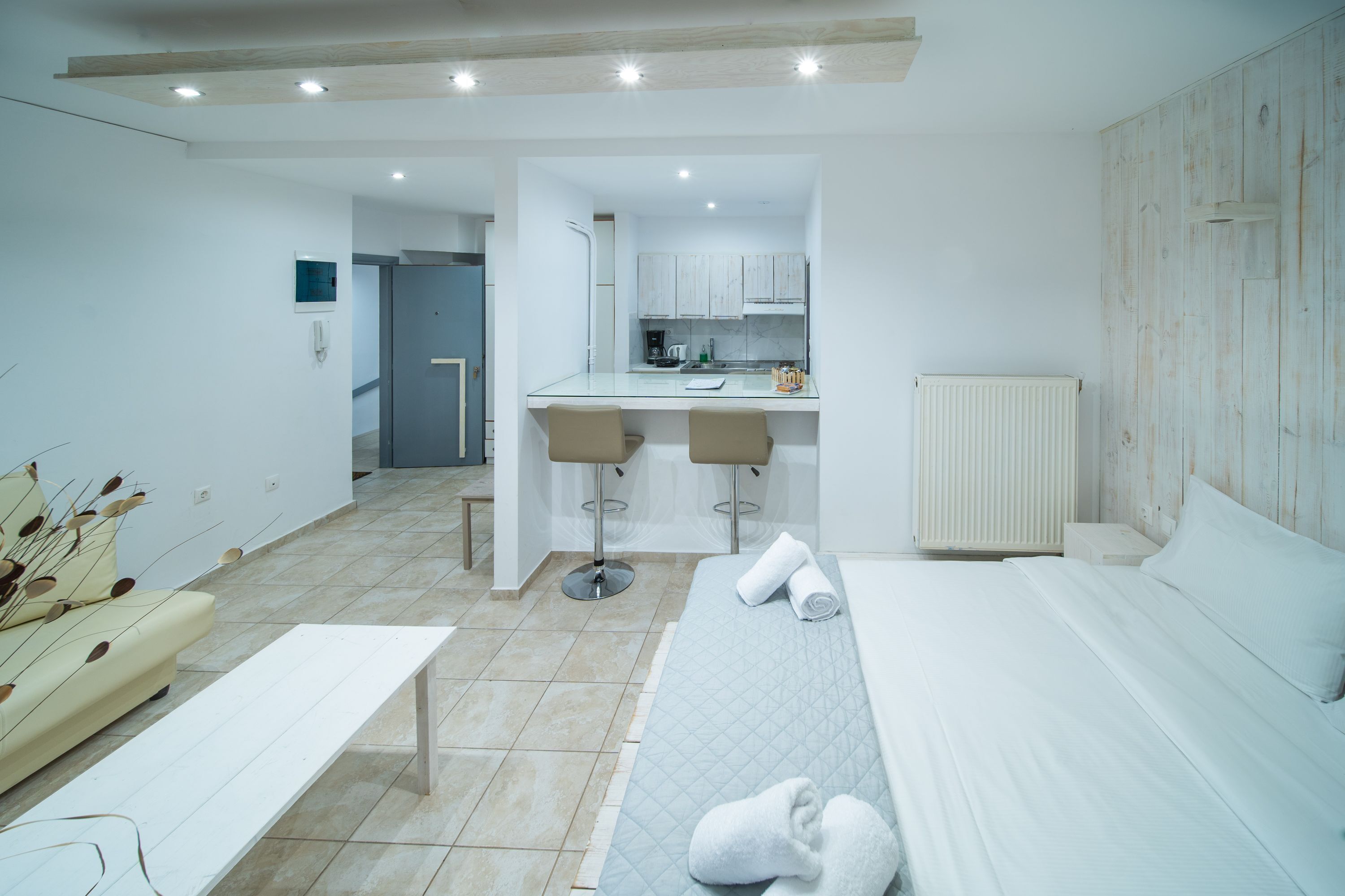A modern hotel room with a bed, a bench, a small table, and a kitchenette in the background.