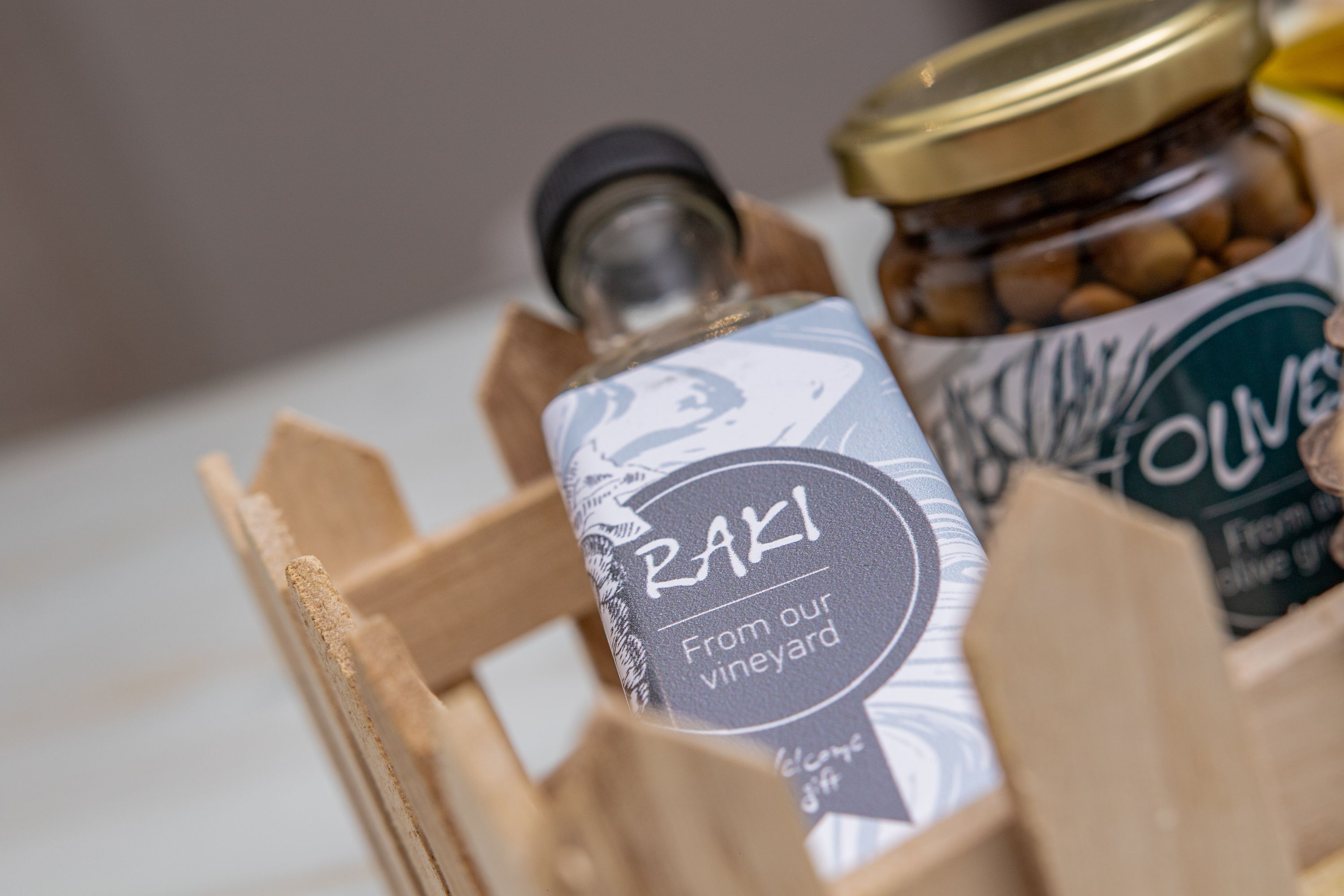 A wooden crate containing a bottle labeled "Raki From our vineyard" and a jar of olives.