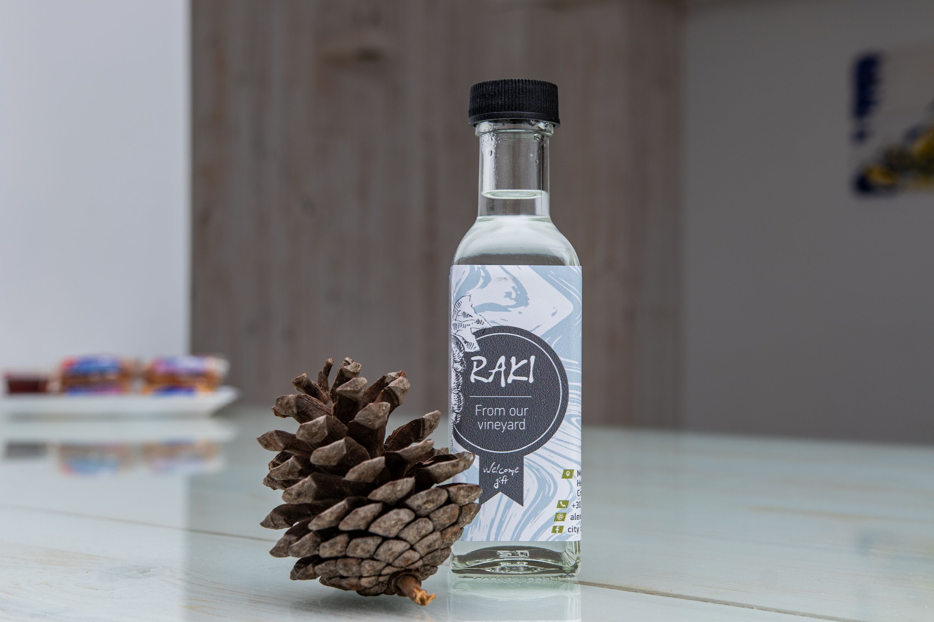 Una bottiglia con un'etichetta che riporta “RAKI Dal nostro vigneto. Omaggio di benvenuto” accanto a una pigna su una superficie riflettente.