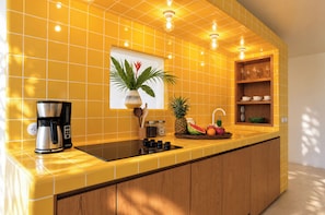 A kitchen with yellow tiles, a coffee maker, a fruit bowl, and a shelf with glasses.