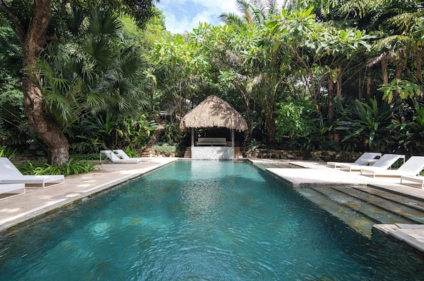 A pool with lounge chairs and a thatched-roof structure surrounded by lush greenery.
