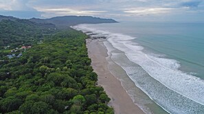 Vista aérea de una zona costera con playa, abundante vegetación y una cadena montañosa distante.