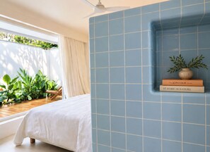 A bedroom with a bed, a window with curtains, a ceiling fan, a blue tiled wall, and a small shelf with books and a plant.