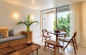 Una sala moderna con sofá, mesa de centro y zona de comedor con sillas. Hay una planta sobre una mesita auxiliar y un ventanal con cortinas.