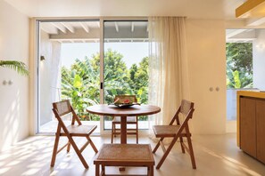 Un comedor con una mesa redonda y cuatro sillas, vistas a exuberante vegetación a través de un ventanal corredero, y un armario de madera a la derecha.