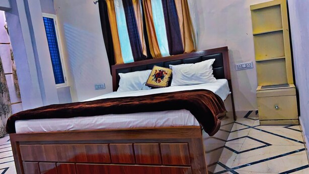 Een net opgemaakt bed met een houten hoofdbord, wit beddengoed en een decoratief kussen. Aan de rechterkant staat een klein nachtkastje met een lamp. De kamer heeft een betegelde vloer met een geometrisch patroon.