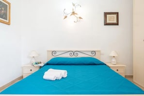 Una cama bien hecha con una colcha azul, dos mesitas de noche con lámparas, y dos cuadros en la pared.