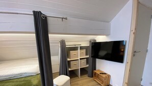 Un dormitorio pequeño con cama, televisor montado en la pared, mueble de almacenamiento con cajones y un taburete de madera.