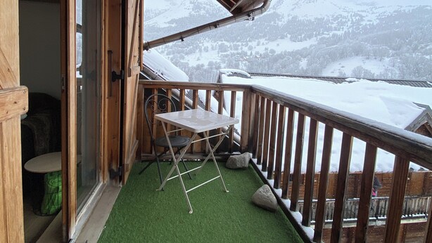 Un balcón de madera con una mesa y una silla, vistas de montañas nevadas y un piso de césped artificial verde.