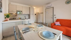 Cocina moderna con armarios blancos, un microondas y un lavabo. Hay una mesa de comedor con platos y vasos. Se ve un sofá naranja al fondo.