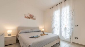Un dormitorio con cama, mesitas de noche, una lámpara de noche, una obra de arte colgada en la pared y una ventana con cortinas estampadas.
