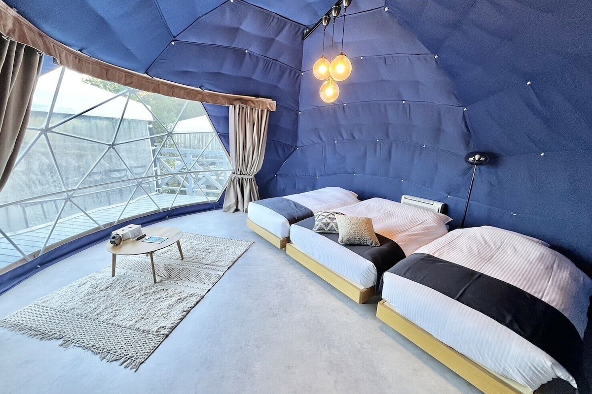 A cozy, modern bedroom with a geometric ceiling design, a large window, and a small table with a lamp.