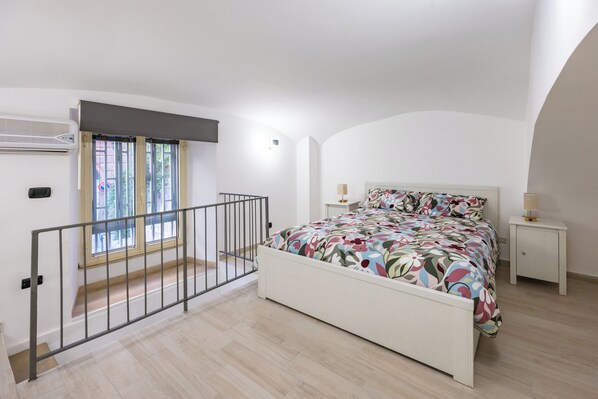 A bedroom with a bed, a nightstand, a lamp, a window with blinds, and a staircase.