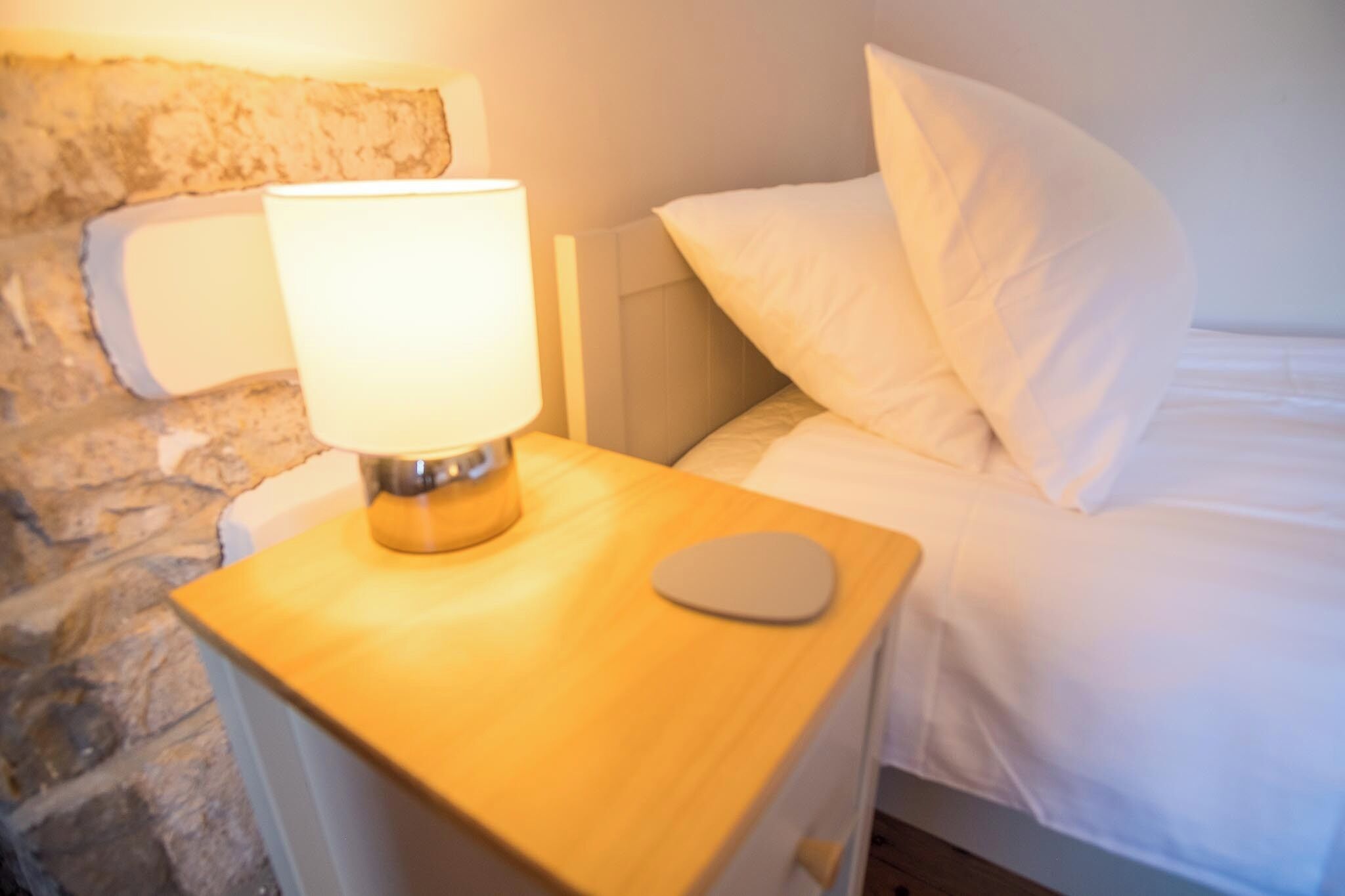 Uma cama com roupa de cama e almofadas brancas, uma mesa de cabeceira com abajur e uma parede de pedra ao fundo.