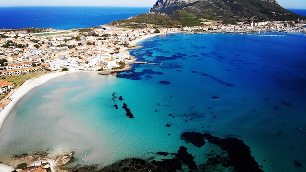 Vista aérea de un pueblo costero con una montaña destacada, aguas azules cristalinas y una zona de playa.
