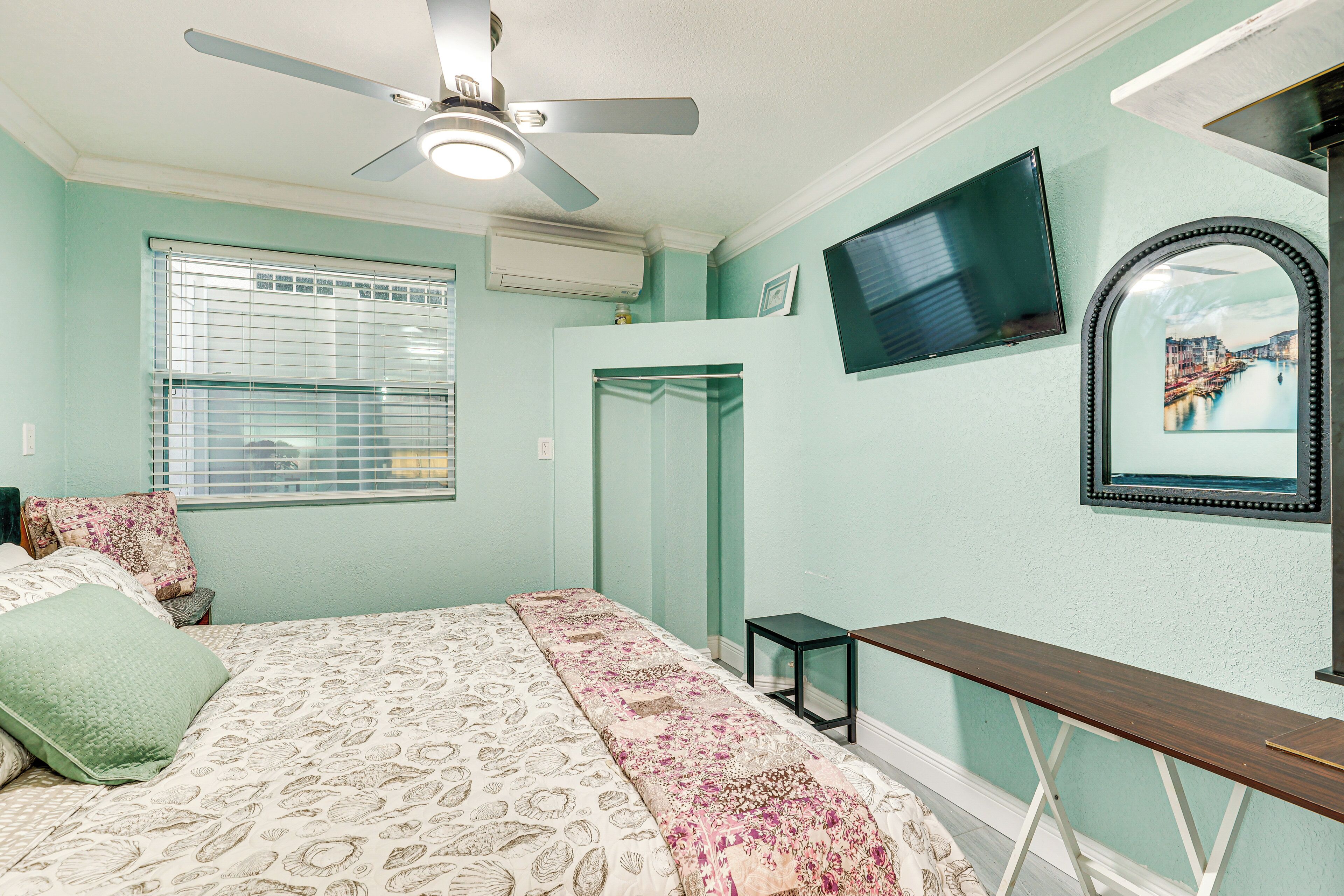 A bedroom with a bed, a ceiling fan, a TV, a mirror, and a window with blinds.