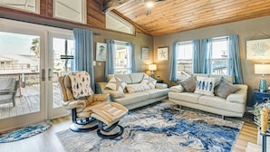 A spacious living room with a large window, a ceiling fan, and a leather chair. - Fish, Crab, Unwind! Canal-front Gem in Galveston (Galveston)
