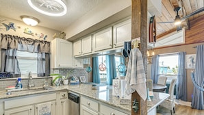 A kitchen with white cabinets, a dishwasher, and a dining area with a table and chairs. - Fish, Crab, Unwind! Canal-front Gem in Galveston (Galveston)