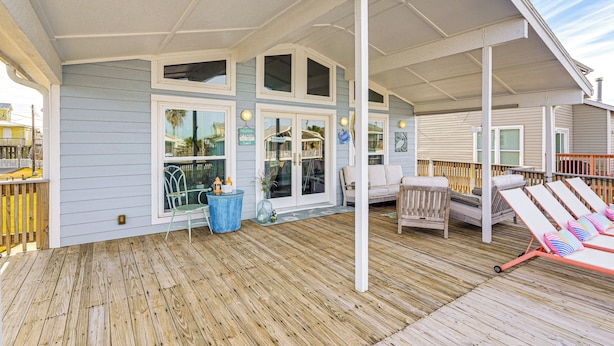 A covered deck with a wooden floor, a white railing, and a seating area with a sofa and lounge chairs. - Fish, Crab, Unwind! Canal-front Gem in Galveston (Galveston)