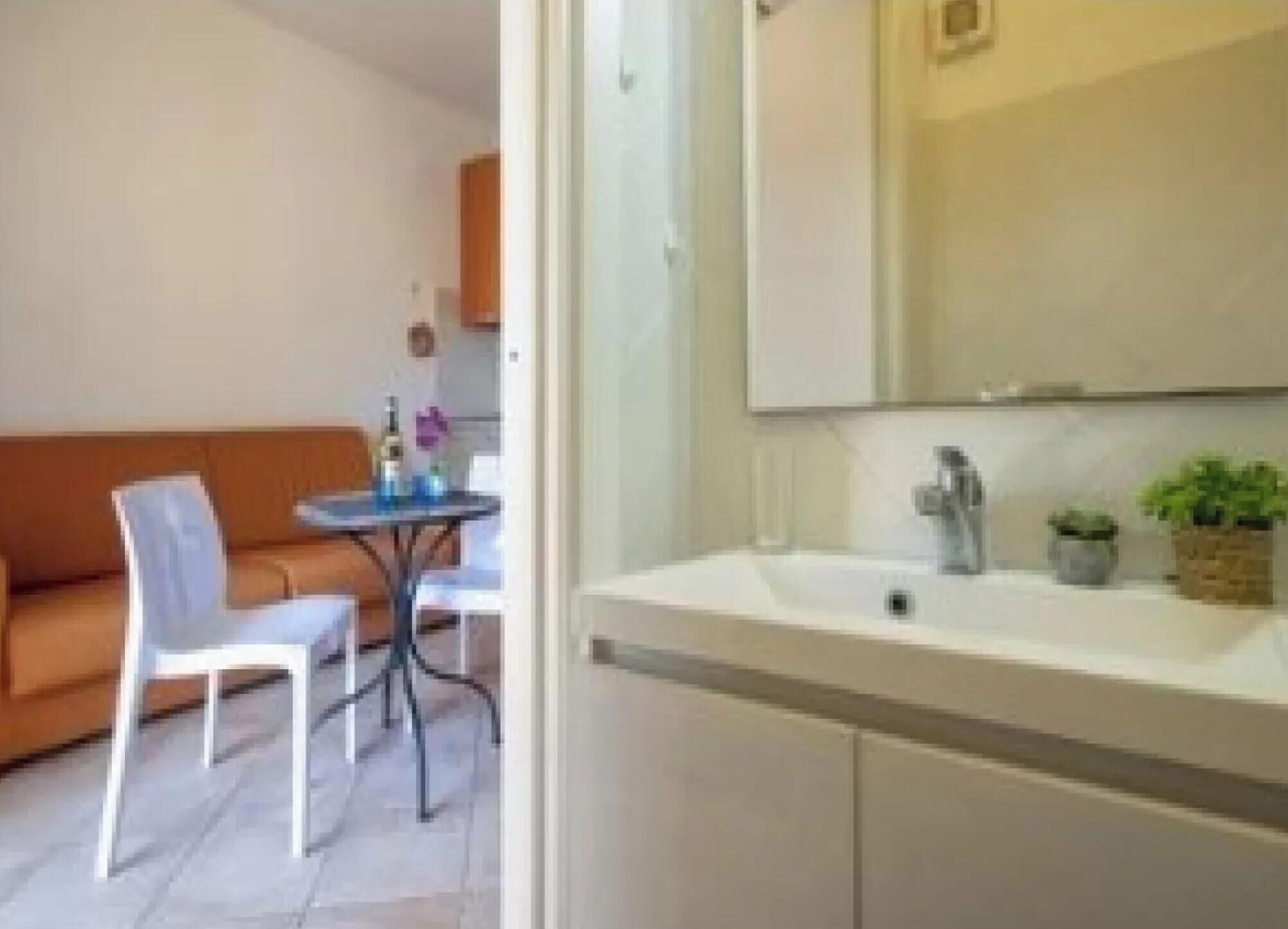 Una stanza piccola e moderna con un divano bianco, un tavolino con una sedia, un lavandino bianco con specchio e due piante in vaso.
