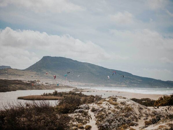 A coastal landscape with a beach, dunes, and a hill in the background.