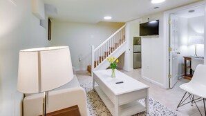 A modern living room with a white sofa, a coffee table with a vase of yellow flowers, a lamp, and a staircase leading to a kitchen area with a TV. - 2 Mi to Dtwn DC: Wfh-friendly Apt (Washington)
