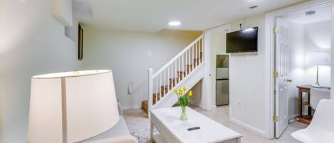 A modern living room with a white sofa, a coffee table with a vase of yellow flowers, a lamp, and a staircase leading to a kitchen area with a TV.