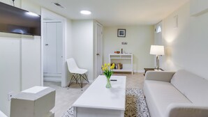 A modern living room with a white sofa, a coffee table with a vase of flowers, a white armchair, a TV mounted on the wall, and a bookshelf with books and decorative items. - 2 Mi to Dtwn DC: Wfh-friendly Apt (Washington)