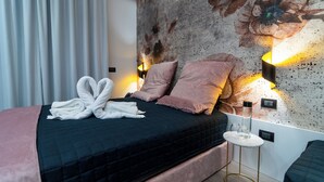 A neatly made bed with a dark quilt, pink pillows, and a pair of white towels folded on top. There is a small round table with a lamp beside the bed. - Luxury Ischia in Naples station train (Napoli)