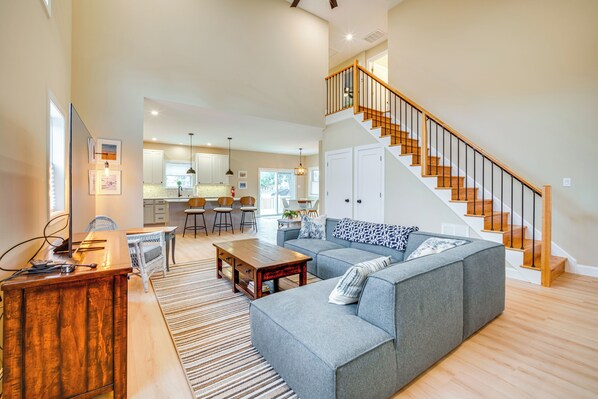 A spacious living room with a grey sofa, wooden coffee table, and a staircase with wooden railings leading to an upper floor. - Steps to Sand: Villas Beach House w/ Decks! (Villas)