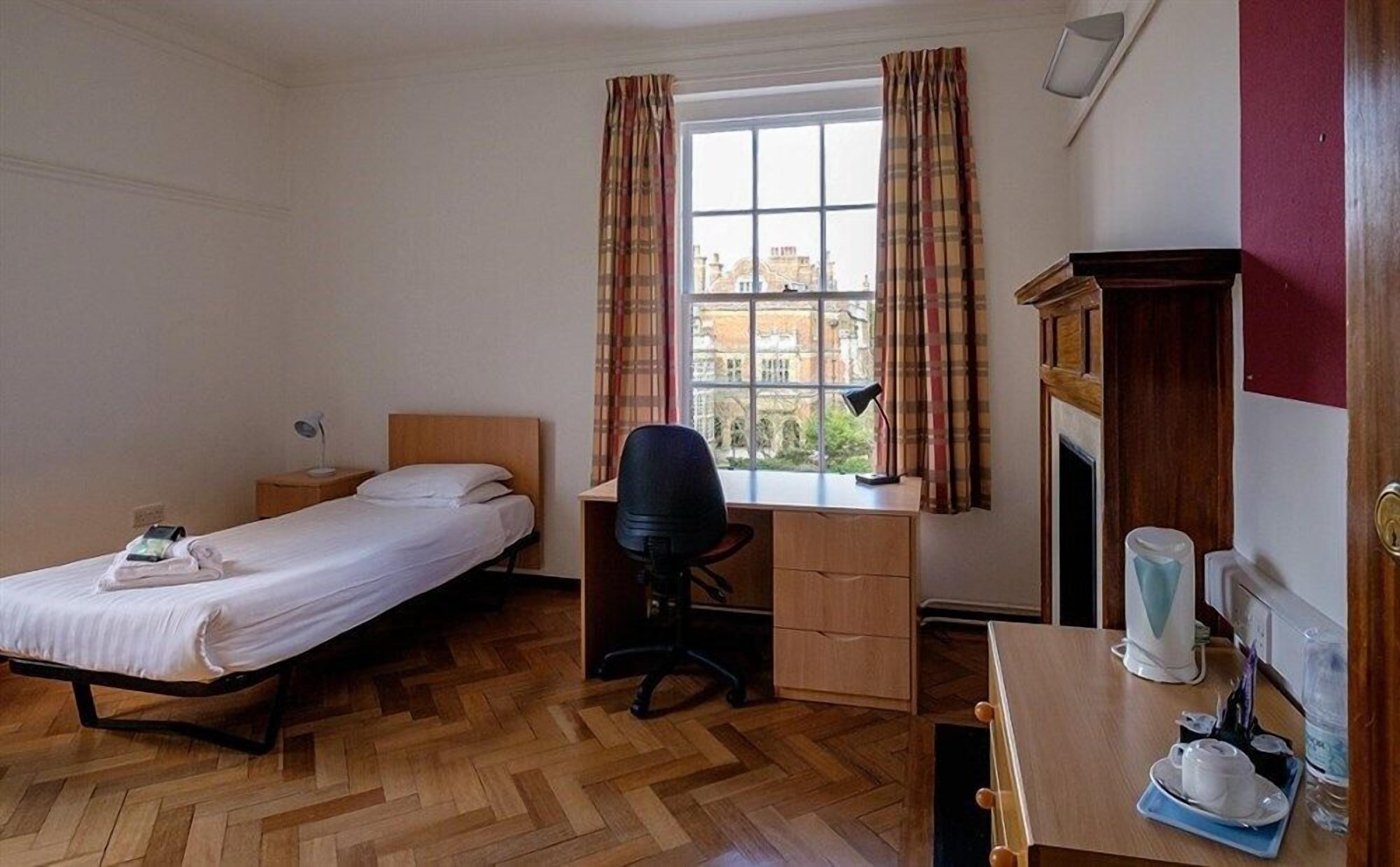 A bedroom with a bed, desk, chair, and a view of buildings through a window.