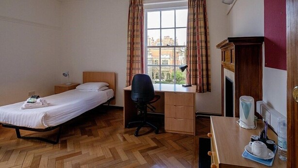 A bedroom with a bed, desk, chair, and a view of buildings through a window. - Sidney Sussex College Cambridge (Cambridge)