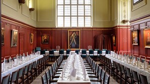 Een formele eetzaal met lange tafels gedekt voor een maaltijd, grote ramen en klassieke wanddecoraties.