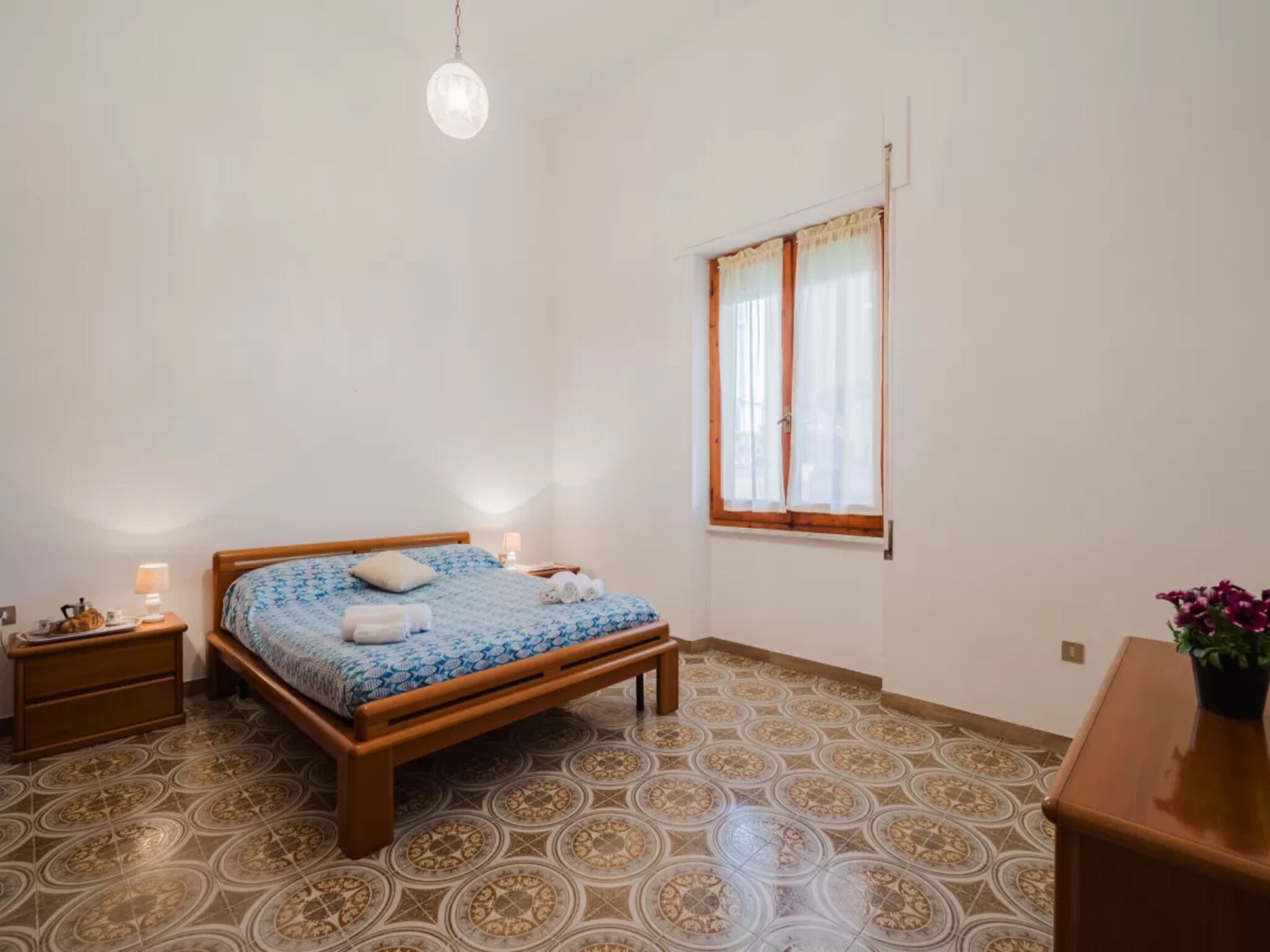 A bedroom with a wooden bed, a nightstand, a lamp, a window with curtains, and patterned floor tiles.