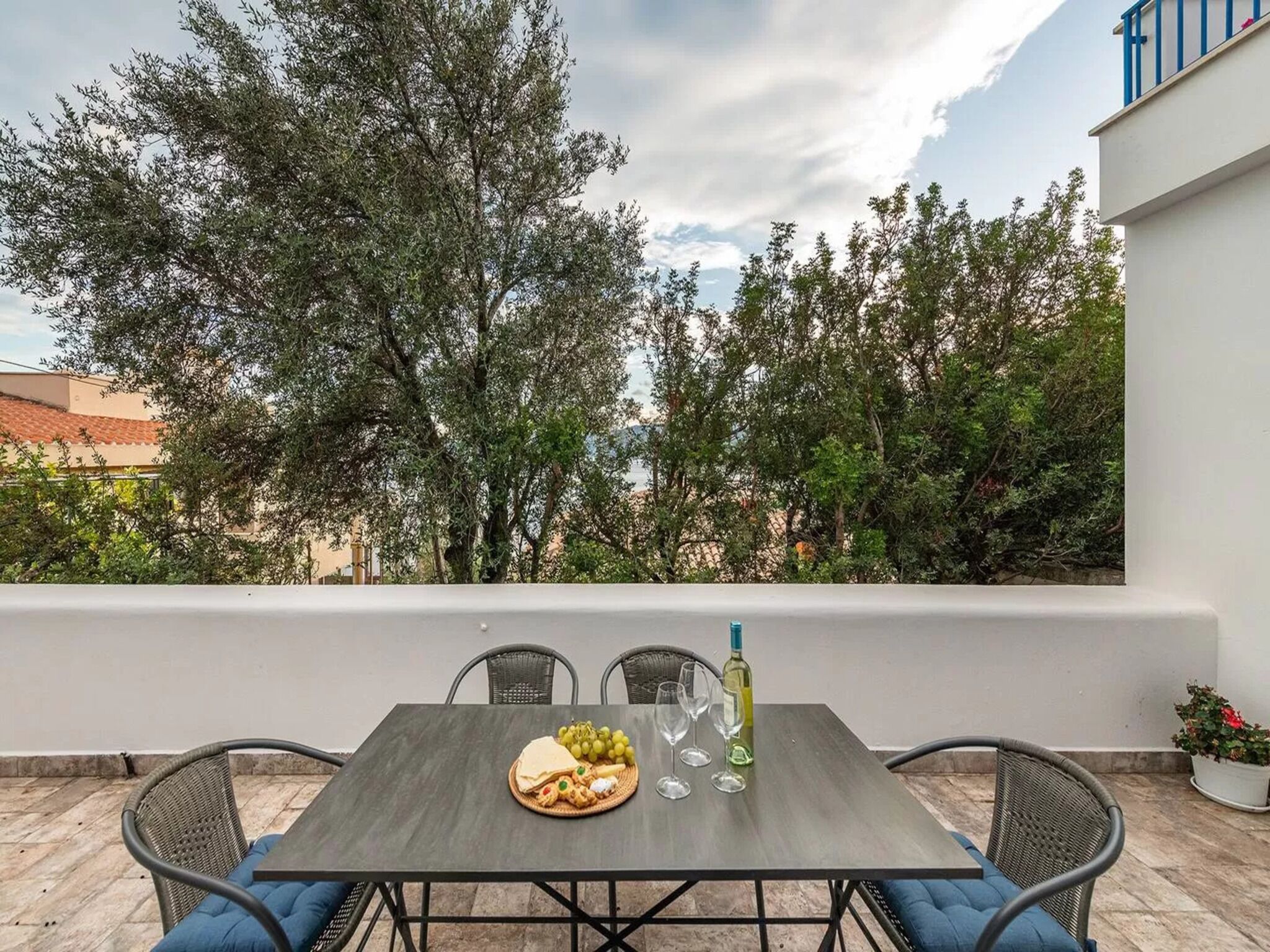 Terrassa exterior amb taula preparada per a dinar, envoltada de cadires, amb vistes a un jardí amb arbres i un edifici al fons.