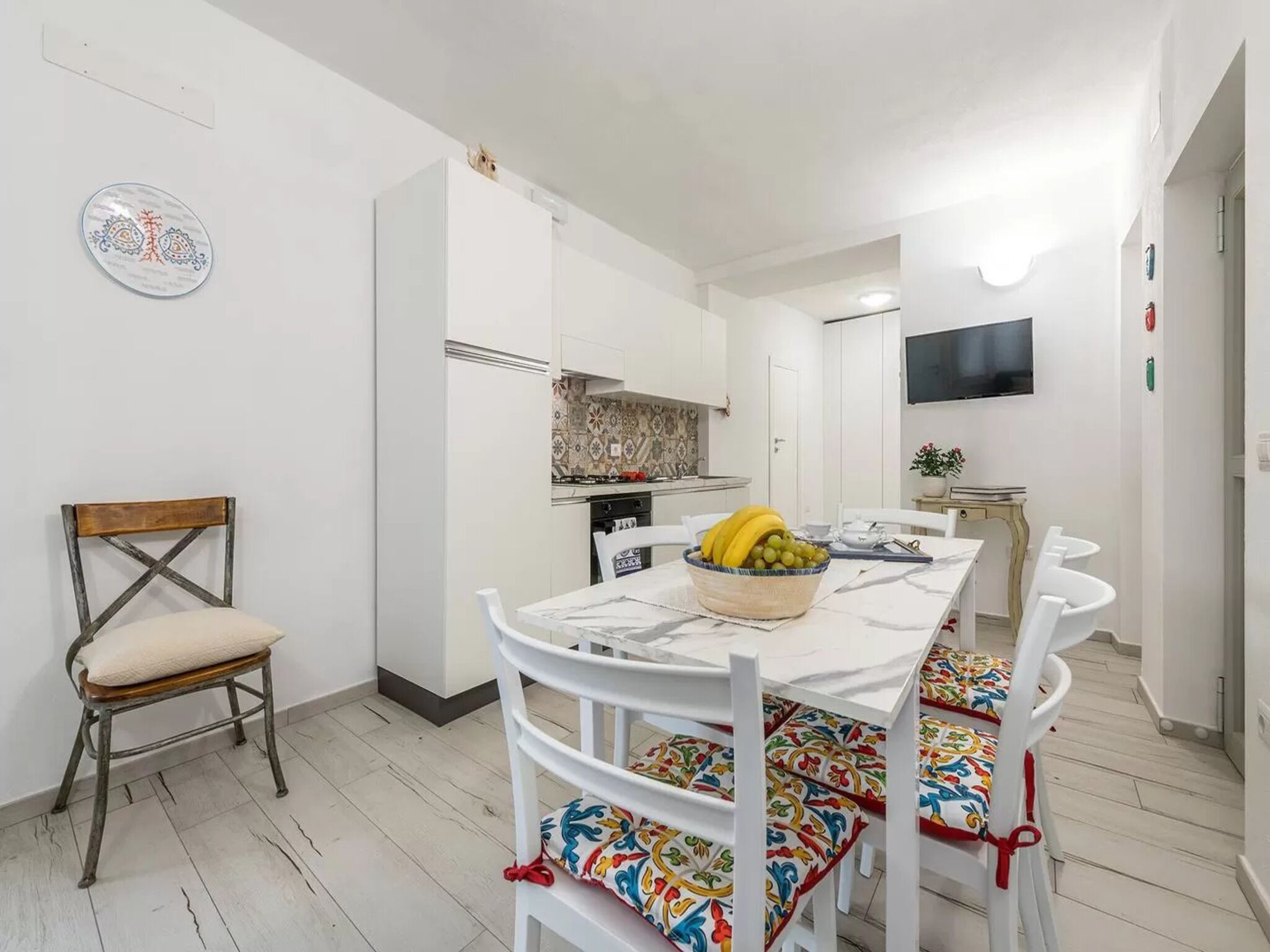 A dining area with a white table, colorful chairs, and a bowl of fruit.