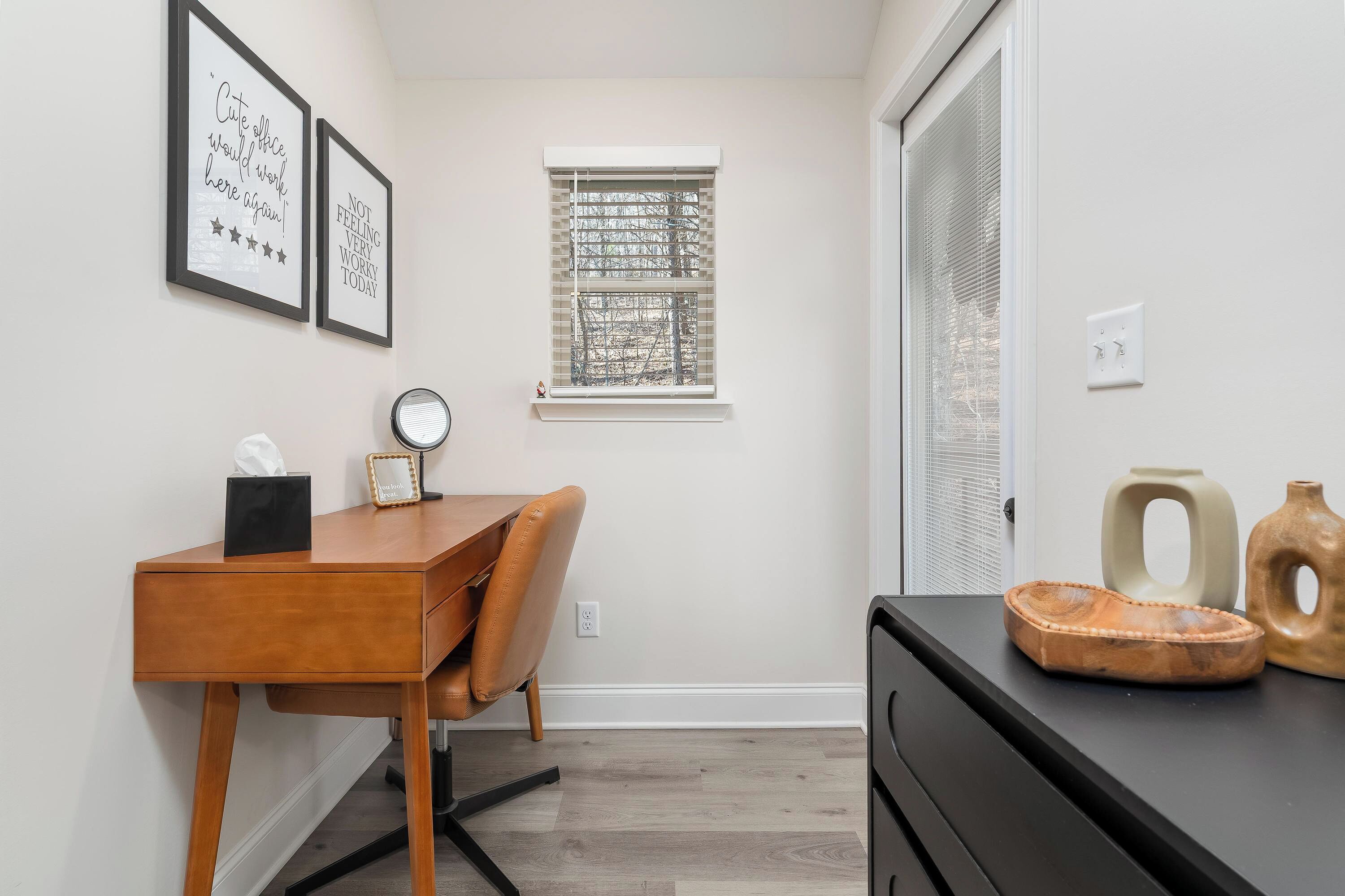 Una habitación pequeña y luminosa con un escritorio de madera, una silla, una ventana con persianas y un aparador negro decorado con objetos.