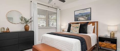 Quarto com cama, ventoinha no teto, mesa de cabeceira, cômoda, janela com cortinas e um quadro na parede.