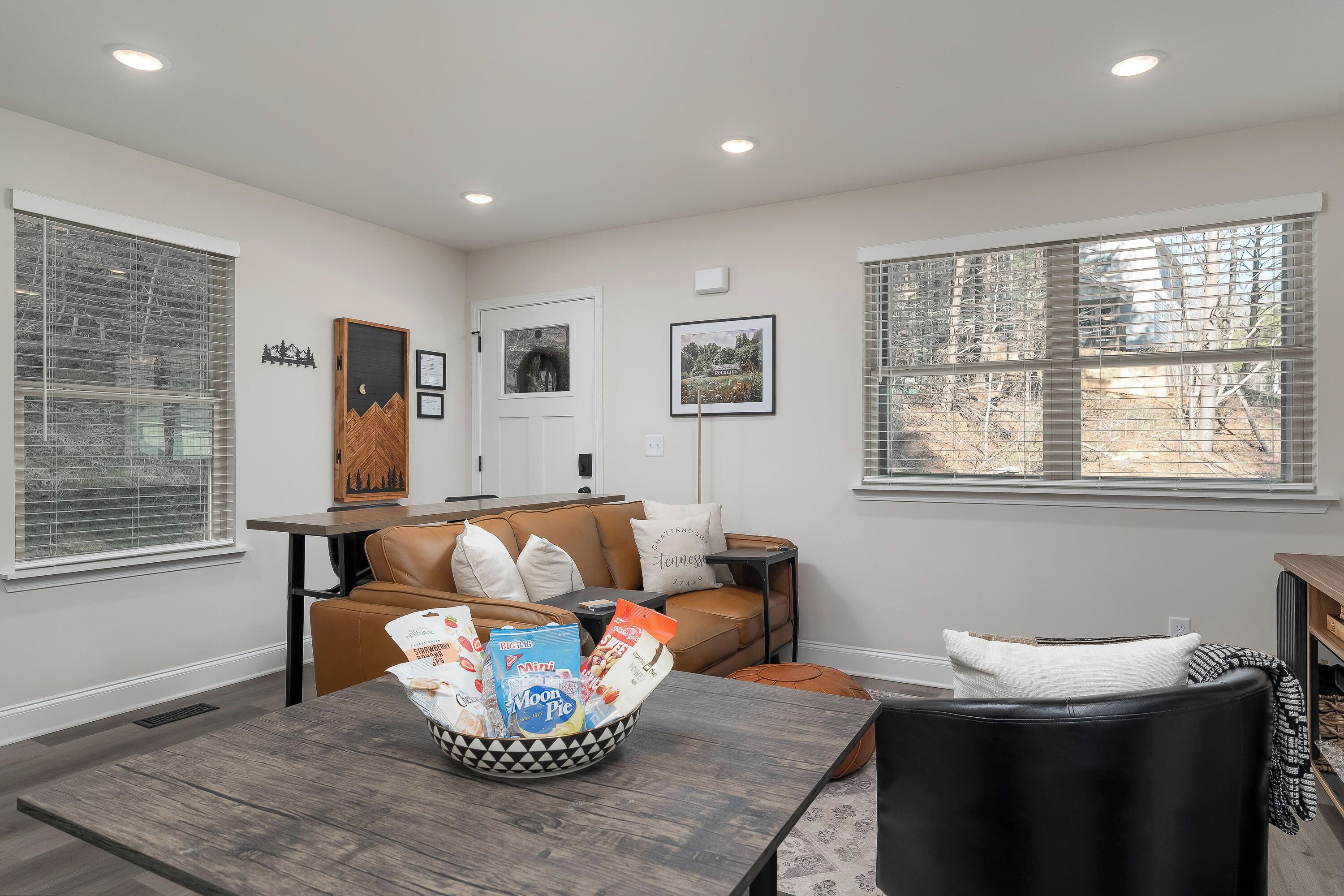 A modern living room with a brown leather sofa, a dining table with a decorative bowl, and large windows with blinds.