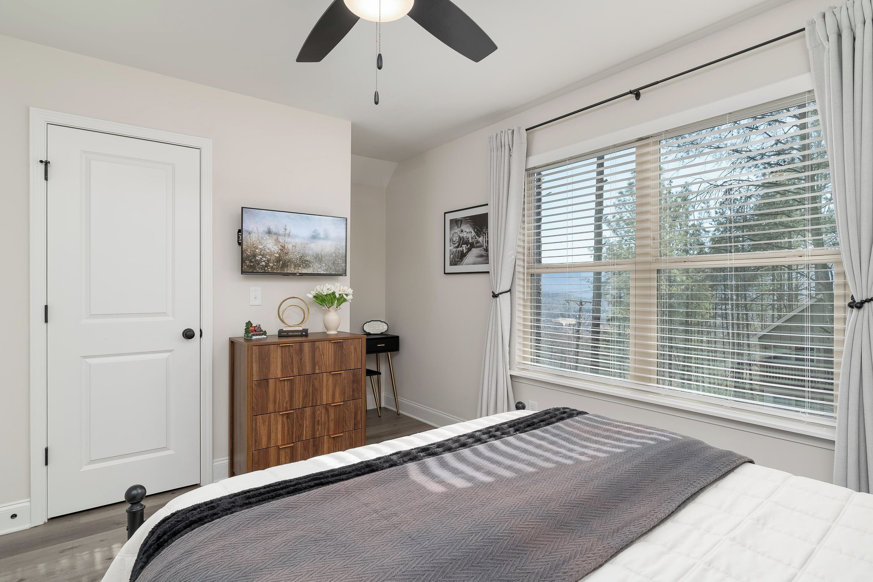 A bedroom with a bed, a ceiling fan, a window with blinds, a desk with a chair, and a door.