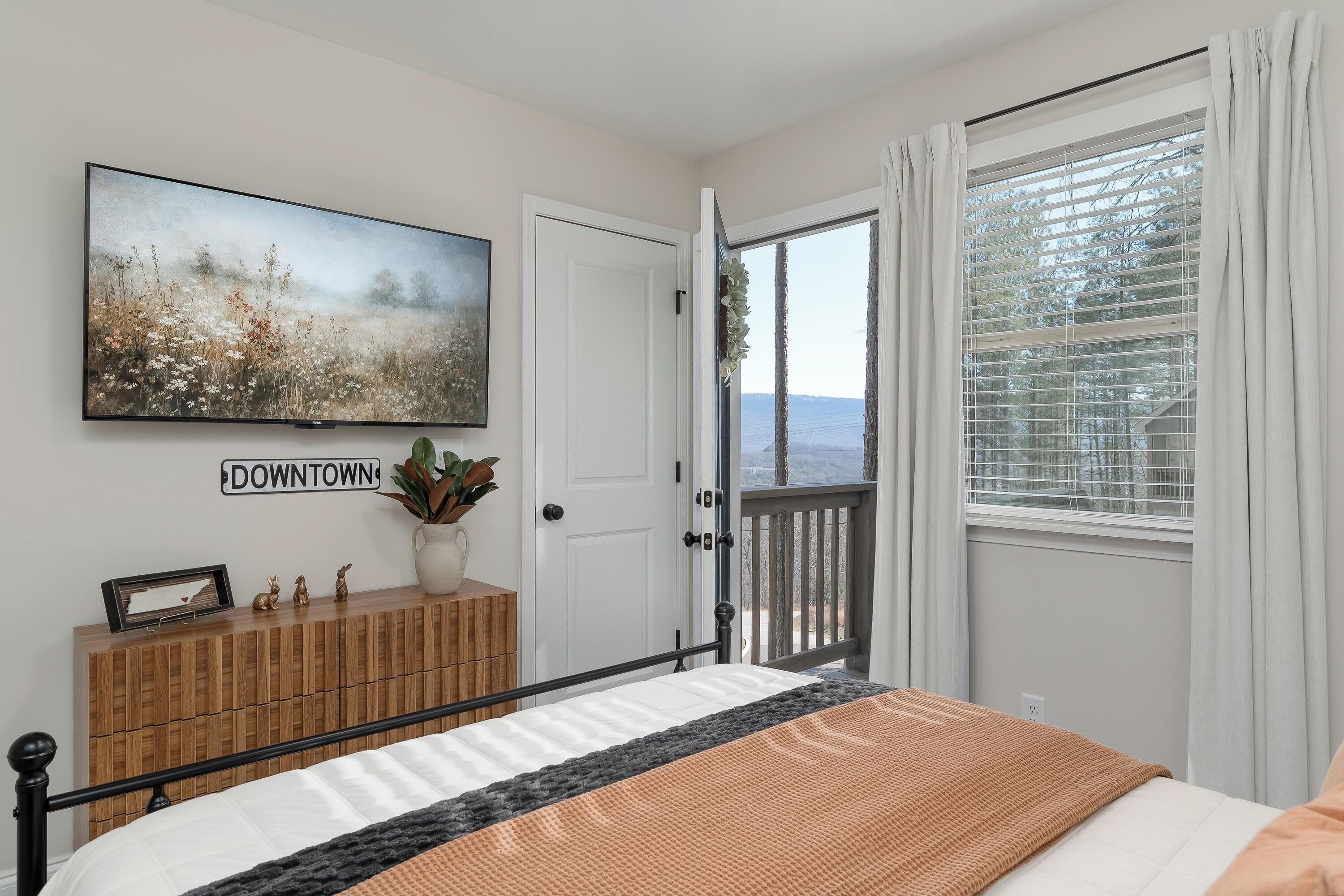 A bedroom with a bed, a dresser, a painting, and a view of the outdoors.