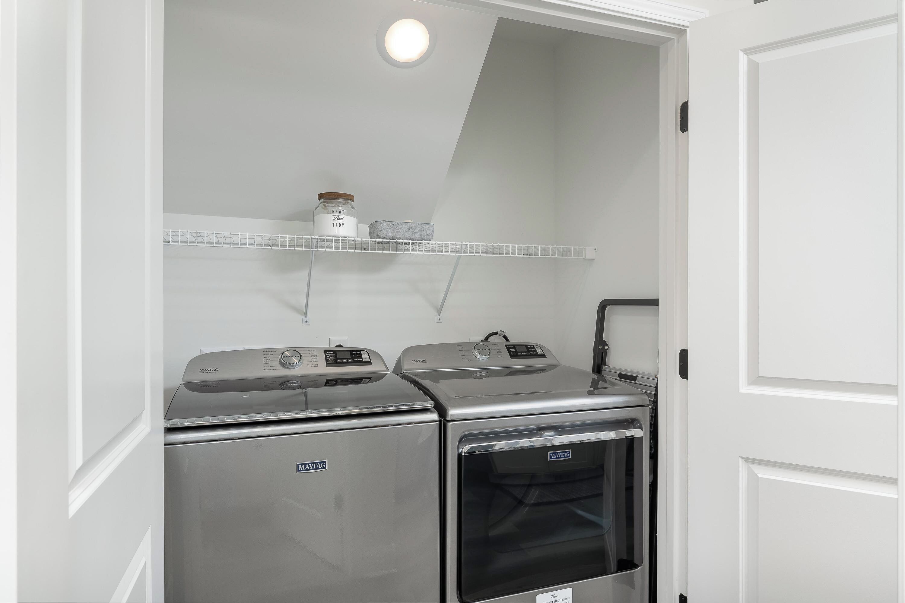 Un lavadero pequeño con dos lavadoras y secadoras Maytag, un estante blanco con recipientes de almacenamiento y una puerta blanca.