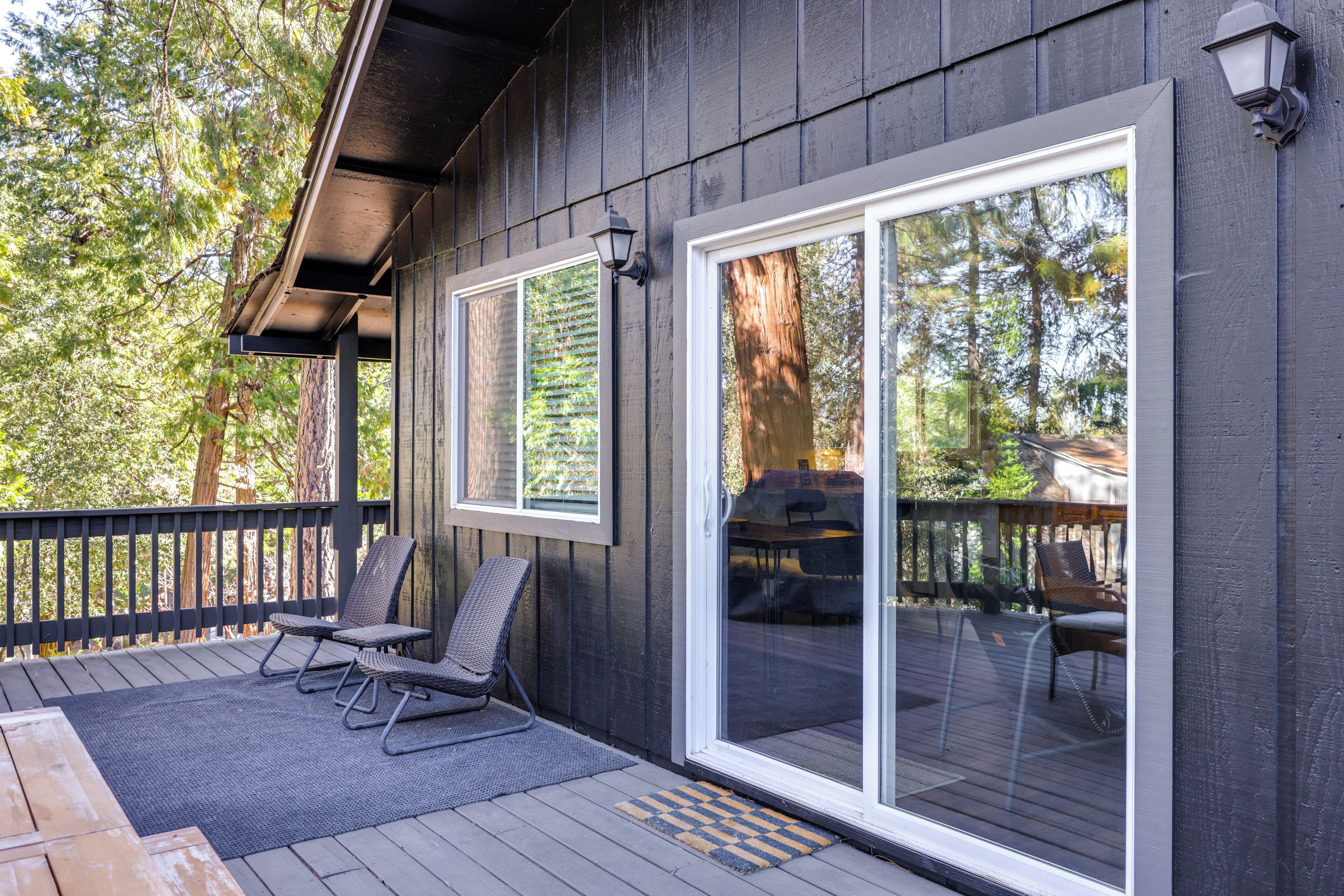 A wooden deck with two chairs and a table, a sliding glass door, and a view of a forest.
