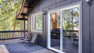 A wooden deck with two chairs and a table, a sliding glass door, and a view of a forest.