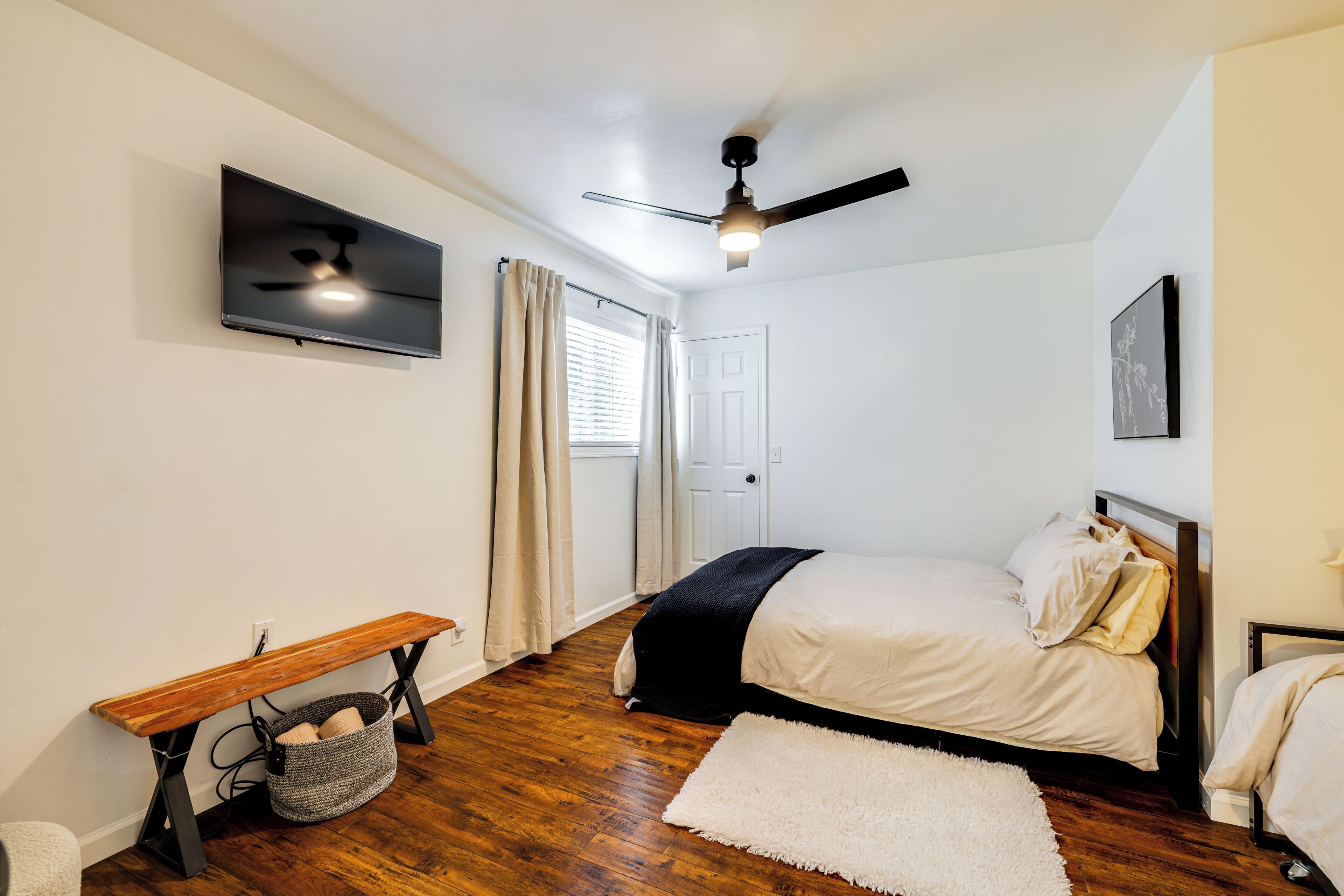 A bedroom with a bed, a bench, a TV, a ceiling fan, and a window with curtains.
