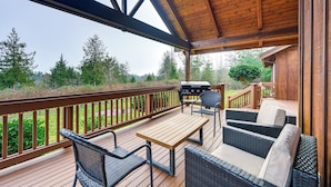 Eine überdachte Terrasse mit Holztisch und Korbmöbeln, mit Blick auf einen Garten und Wald.