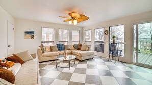 Un salon avec un canapé d’angle beige, une table basse en verre et un ventilateur de plafond. De grandes fenêtres sont équipées de stores, et il y a un balcon avec une table, ainsi qu’un sol à carreaux.