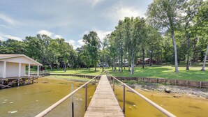 Une jetée en bois qui mène à un quai au-dessus d’une étendue d’eau, avec une zone herbeuse et des arbres en arrière-plan.