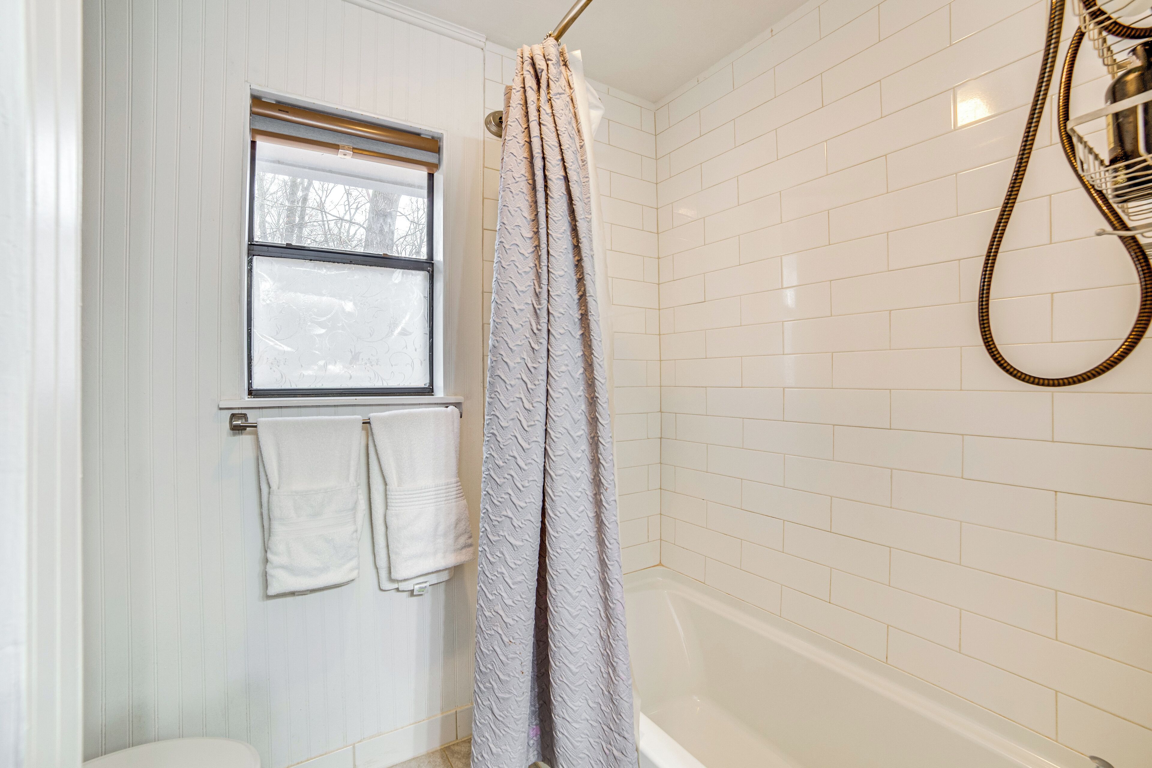 A bathroom with white tiled walls, a bathtub, a shower curtain, and a window with a towel hanging on the door.